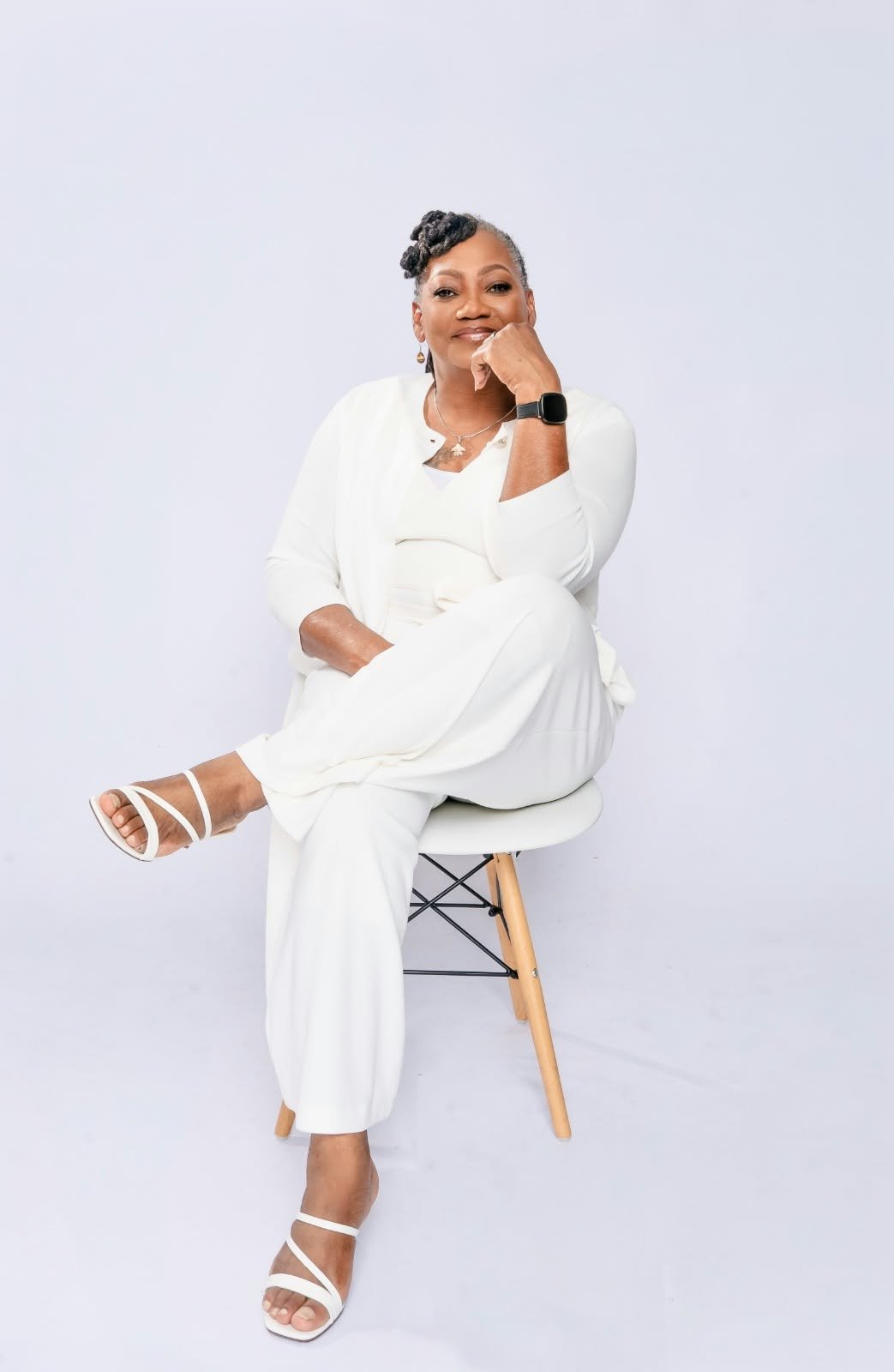 A woman in white seated with her legs crossed and her chin resting on one hand.